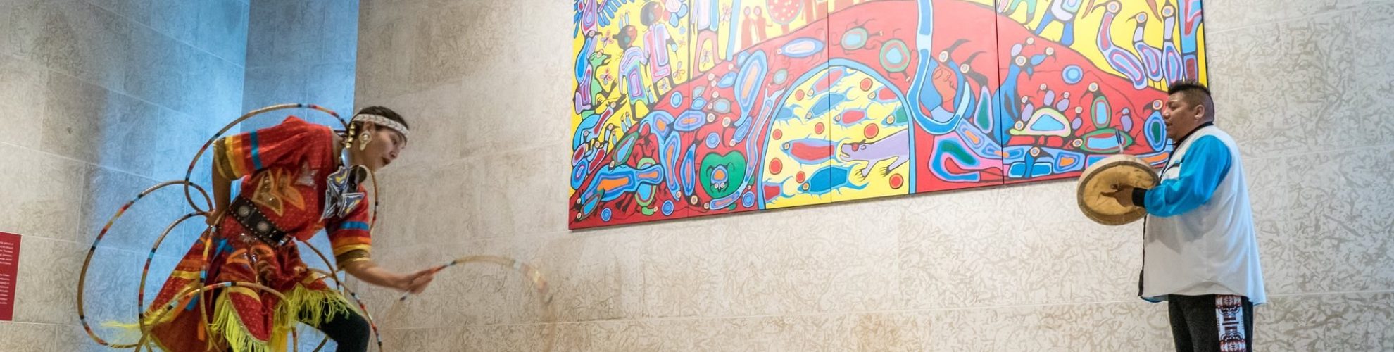 One person hoop dancing and one drumming in front of a mural at the WAG.