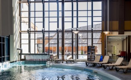 Lounge chairs lined up along the side of a pool in front of a wall of glass windows looking out.