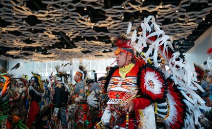 Indigenous person dressed in traditional head dress, dancing with others at Manito Ahbee Festival pow wow.